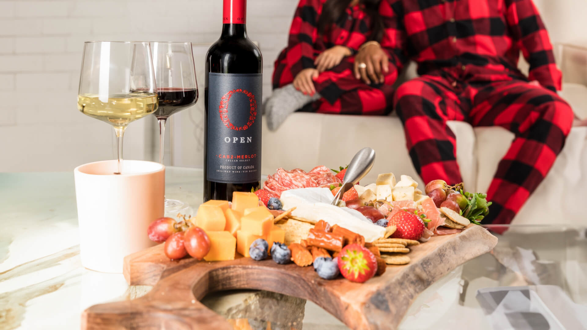 Couple cozied up, in matching plaid pajamas enjoying Open Cab-Merlot wine and a charcuterie board.
